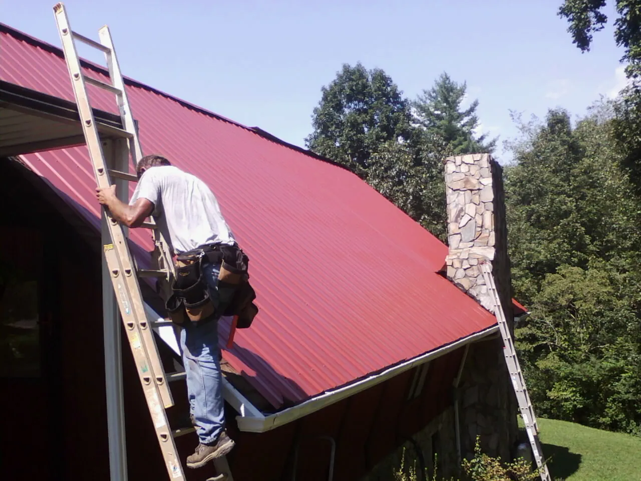 Roofer on a ladder beside a red metal roof and stone chimney — Soffit & Fascia Repair by Roofing Brothers in Cross Hill